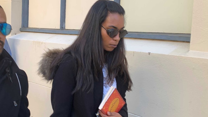 A woman in a black jacket and holding an orange book outside court holding the hand of a man.