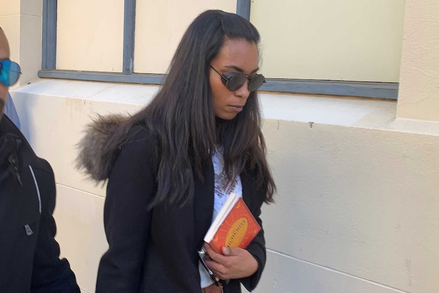 A woman in a black jacket and holding an orange book outside court holding the hand of a man.