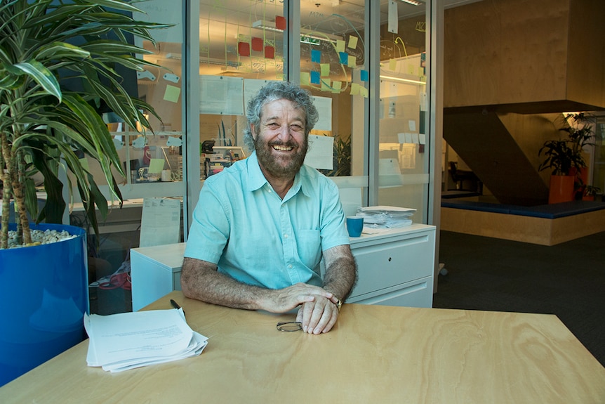 Man in a university office wearing a black shirt smiling from ear to ear.