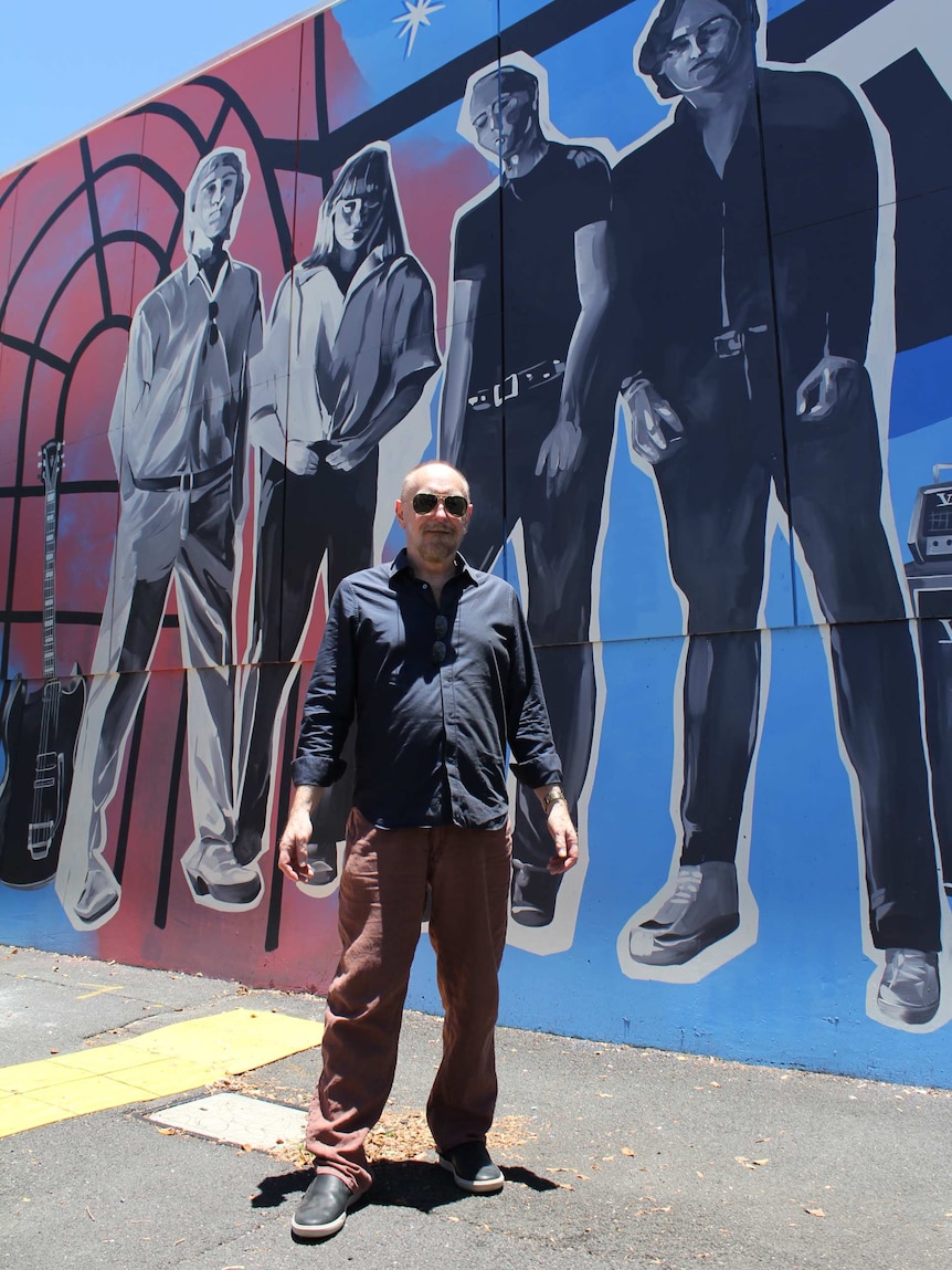 The Saints guitarist Ed Kuepper standing in front of a black, white, blue and red mural of his bandmates.