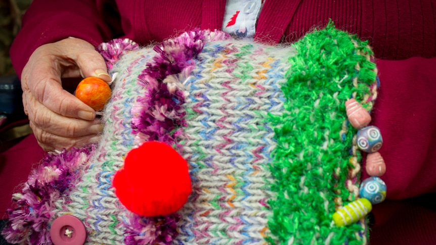 An elderly hand plays with a bead sewn into a brightly coloured knitted fiddle muff.