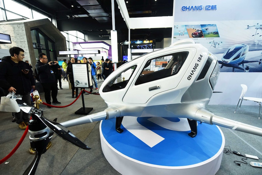 A white passenger drone is on display behind a red rope and a crowd looks at it and takes pictures.