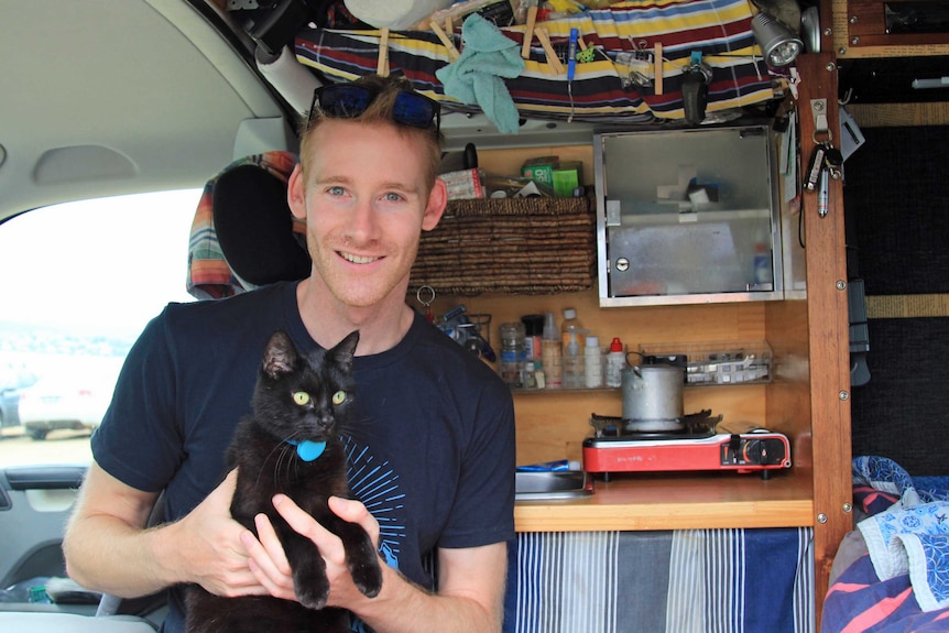 A man smiling and holding a black cat inside a campervan