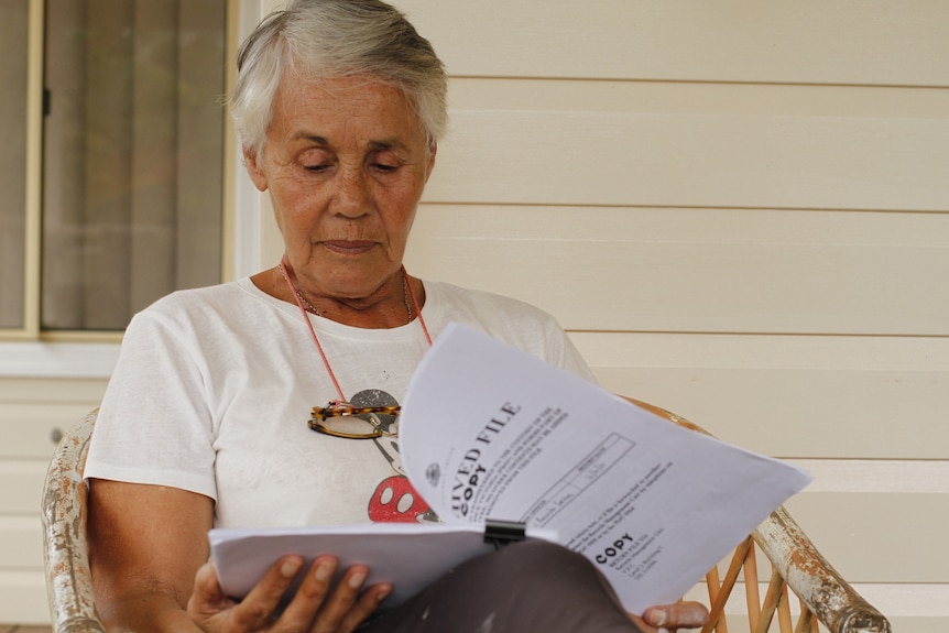 A woman wearing a white t-shirt and glasses sits and reads a document.