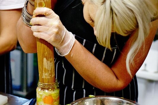 A workshop participant uses a wooden rolling pin to pack down food scraps in a pickling lesson.