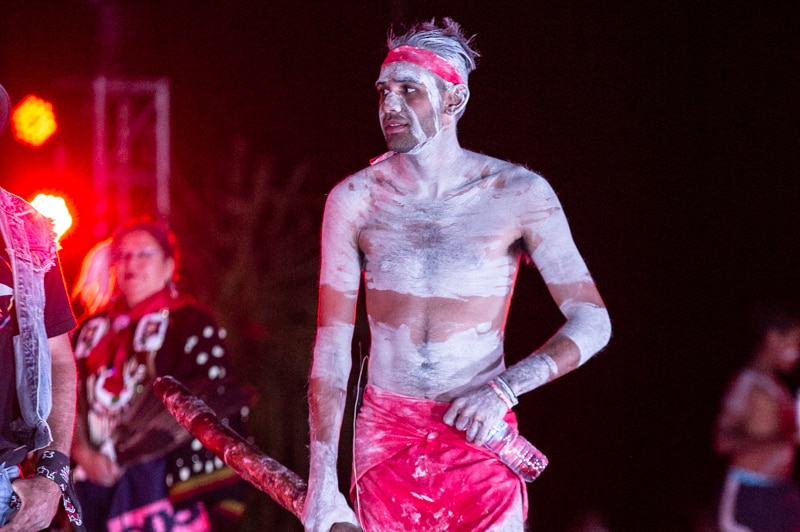 A young Indigenous man covered in traditional body paint holds a didgeridoo.