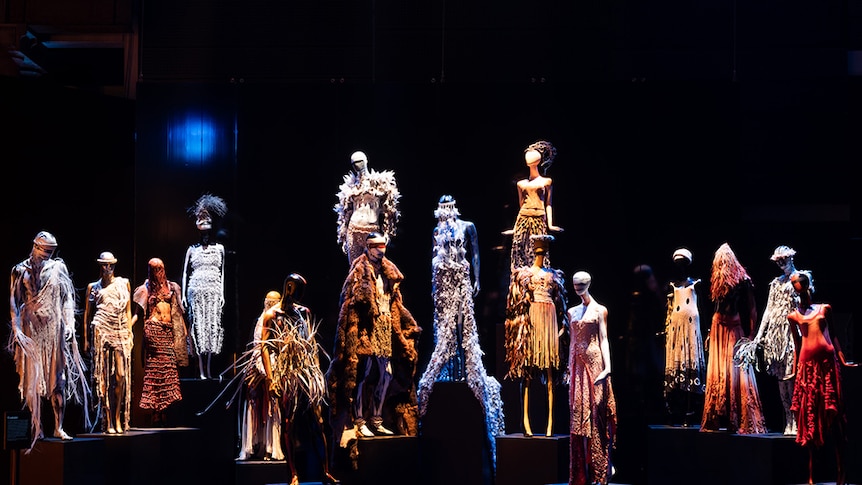 16 staggered mannequins dressed in Bangarra Dance Theatre costumes stand on display in dark exhibition space.
