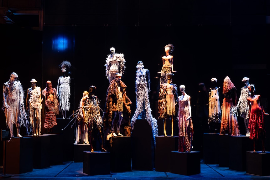 16 staggered mannequins dressed in Bangarra Dance Theatre costumes stand on display in dark exhibition space.