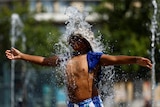 Water from a fountain splashes over a boy's face. He has his arms spread.