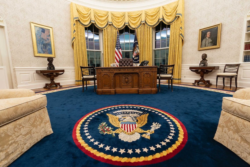 A wide photo of the Oval Office, including a blue rug, gold curtains and the Resolute Desk.