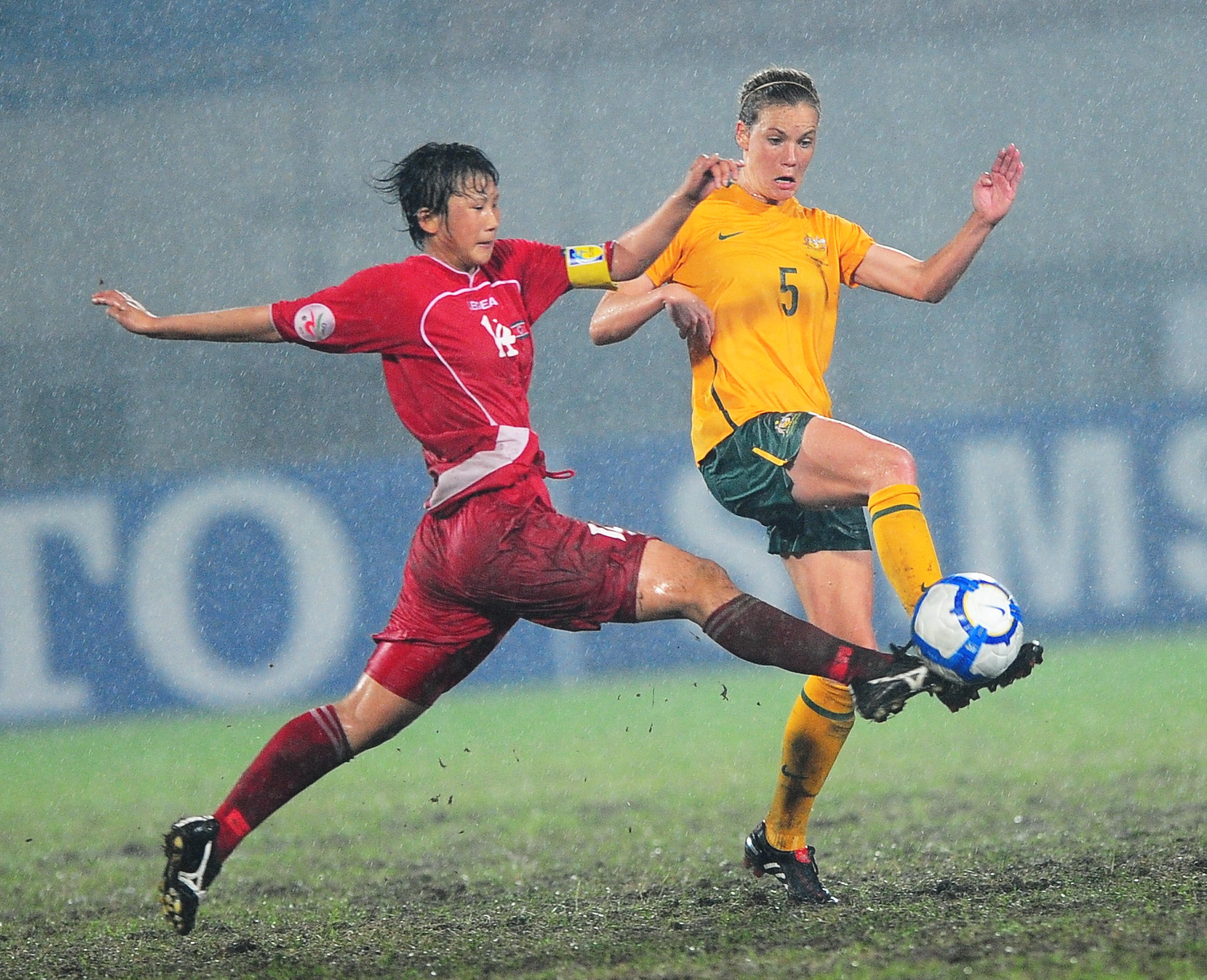 Dois jogadores de futebol, um de amarelo e outro de vermelho, disputam a bola em condições de lama