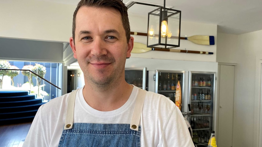 A young male restaurant owner wearing a denim apron standing in front of a bar.