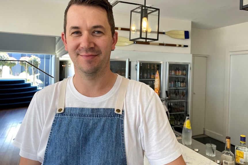 A young male restaurant owner wearing a denim apron standing in front of a bar