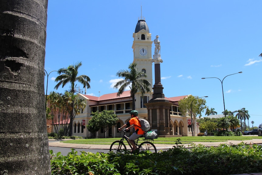 A person in an orange high-vis shirt riding a bike through a roundabout with a clock tower and memorial statue behind