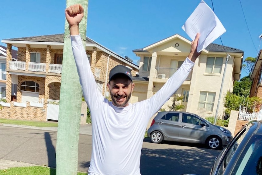 A happy, bearded man in a long-sleeve skivvy and dark blue cap holds his arms outstretched above his head, in a V shape.