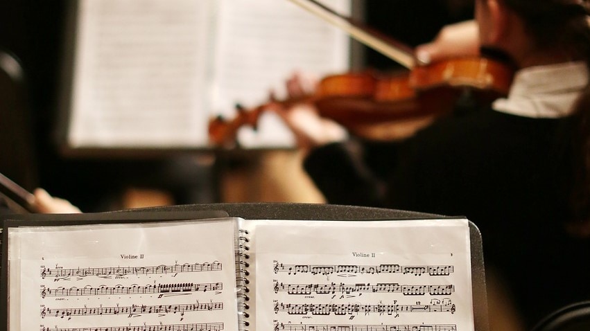 The view from inside an orchestra. Sheet music on a music stand with a violinist playing in the background.