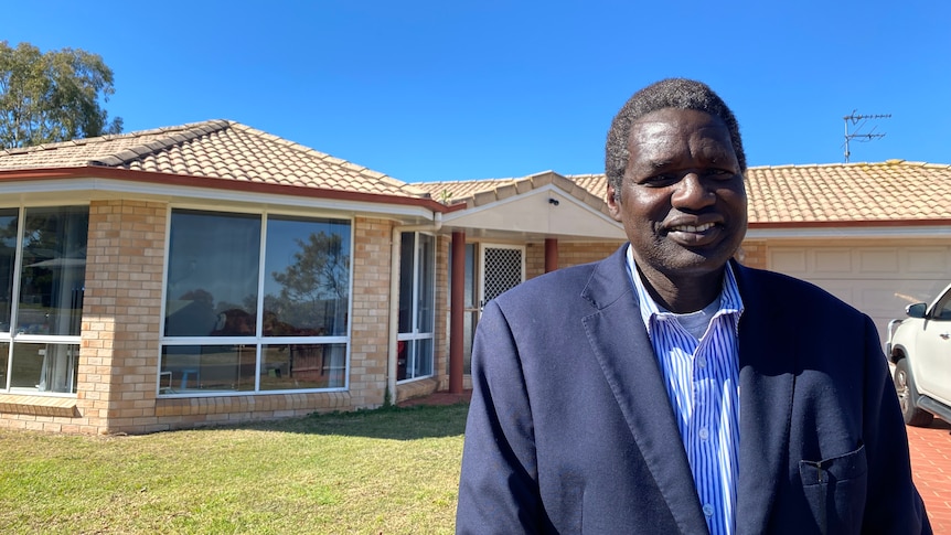 A South Sudanese man standing in front of a brick house. He is wearing a suit and smiling. 