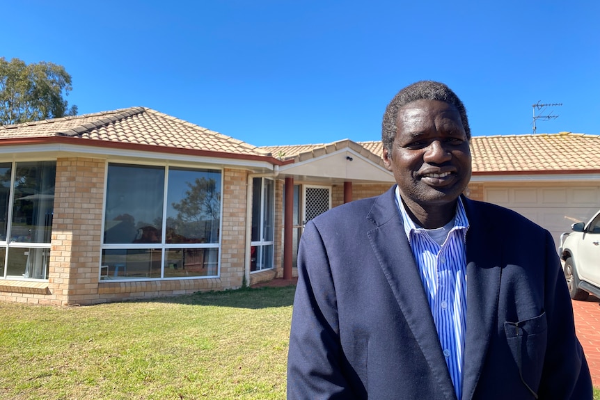 A South Sudanese man standing in front of a brick house. He is wearing a suit and smiling. 