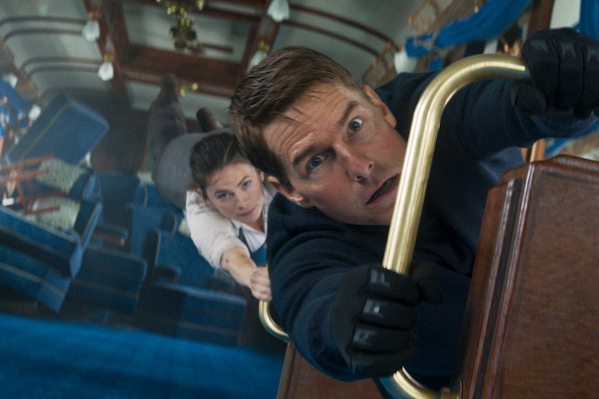 Hayley Atwell, a white brunette woman, and Tom Cruise, a white brunette man, hang onto railings in an upturned train carriage.