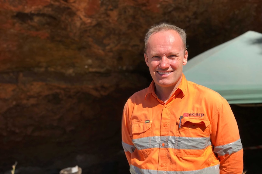 A man stands in front of a rock face, wearing an orange hi-vis uniform