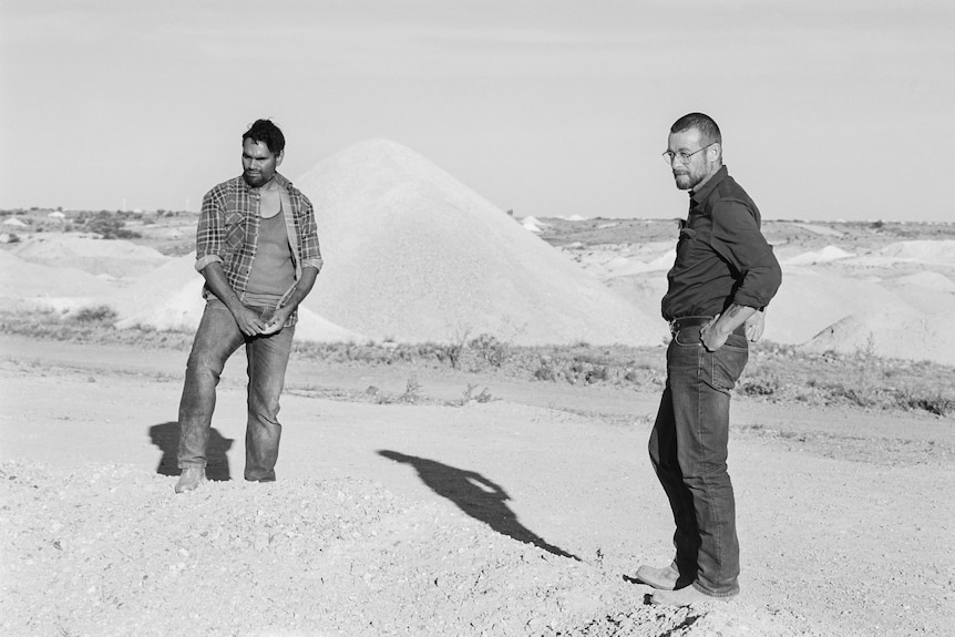 Two men stand in front of a mound of dirt.