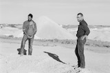 Two men stand in front of a mound of dirt.