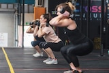Three women in a gym, squatting while holding dumbbells. In between each woman is electrical tape marking physical distancing.