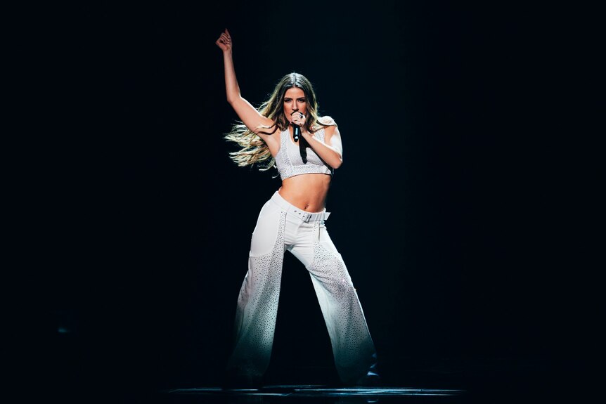 Silia Kapsis with arm raised and a white crop top and white pants, holding microphone, long hair