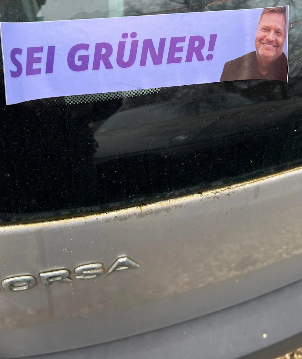 A sticker which says 'sei gruner!" or be greener! in english is stuck on a car with a smiling man next to the slogan
