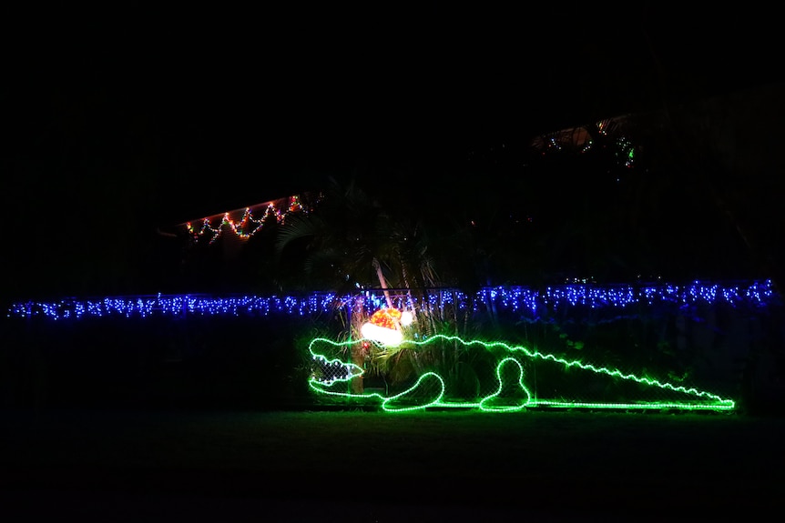 An outline of a crocodile made with green lights hung on a fence, it has a Santa hat also made of lights.