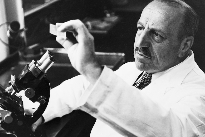 A man in a white lab coat, with a microscope, holding up a glass slide