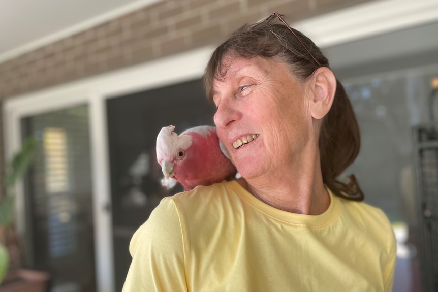 A woman, smiling, in a yellow t-shirt with a pink and white galah sitting on her shoulder.