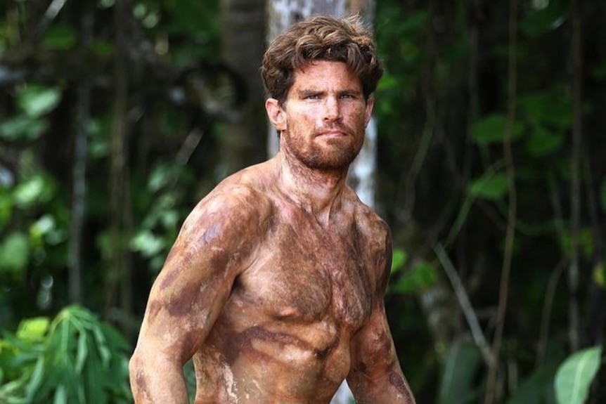 A shirtless man covered in paint stands in front of jungle.