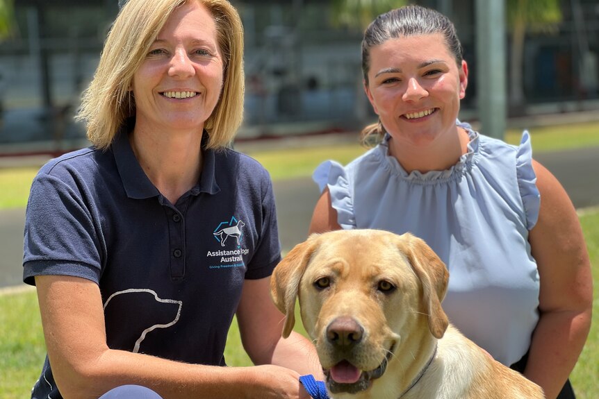 Two women, one blonde wears blue t-shirt with Assistance Dogs Australia logo, other wearing sleeveless powder blue top hold dog.