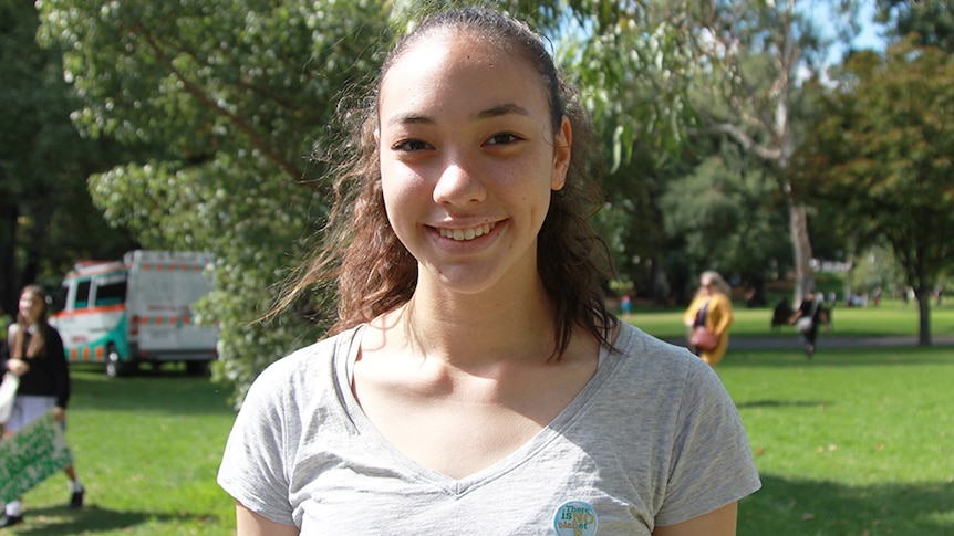 A teenage girl in a grey T-shirt stands in a sunny park.