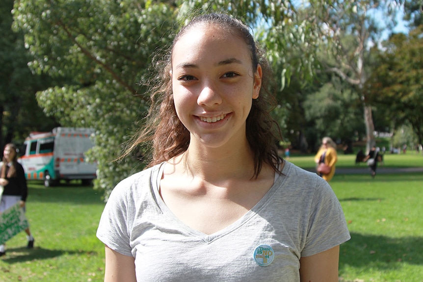 A teenage girl in a grey T-shirt stands in a sunny park.