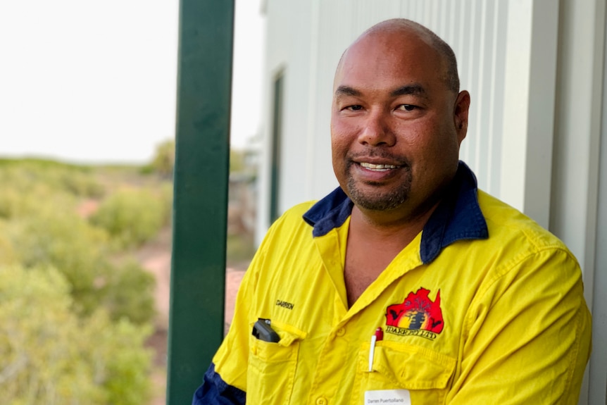 A man in a yellow work shirt stands in front of a shed.