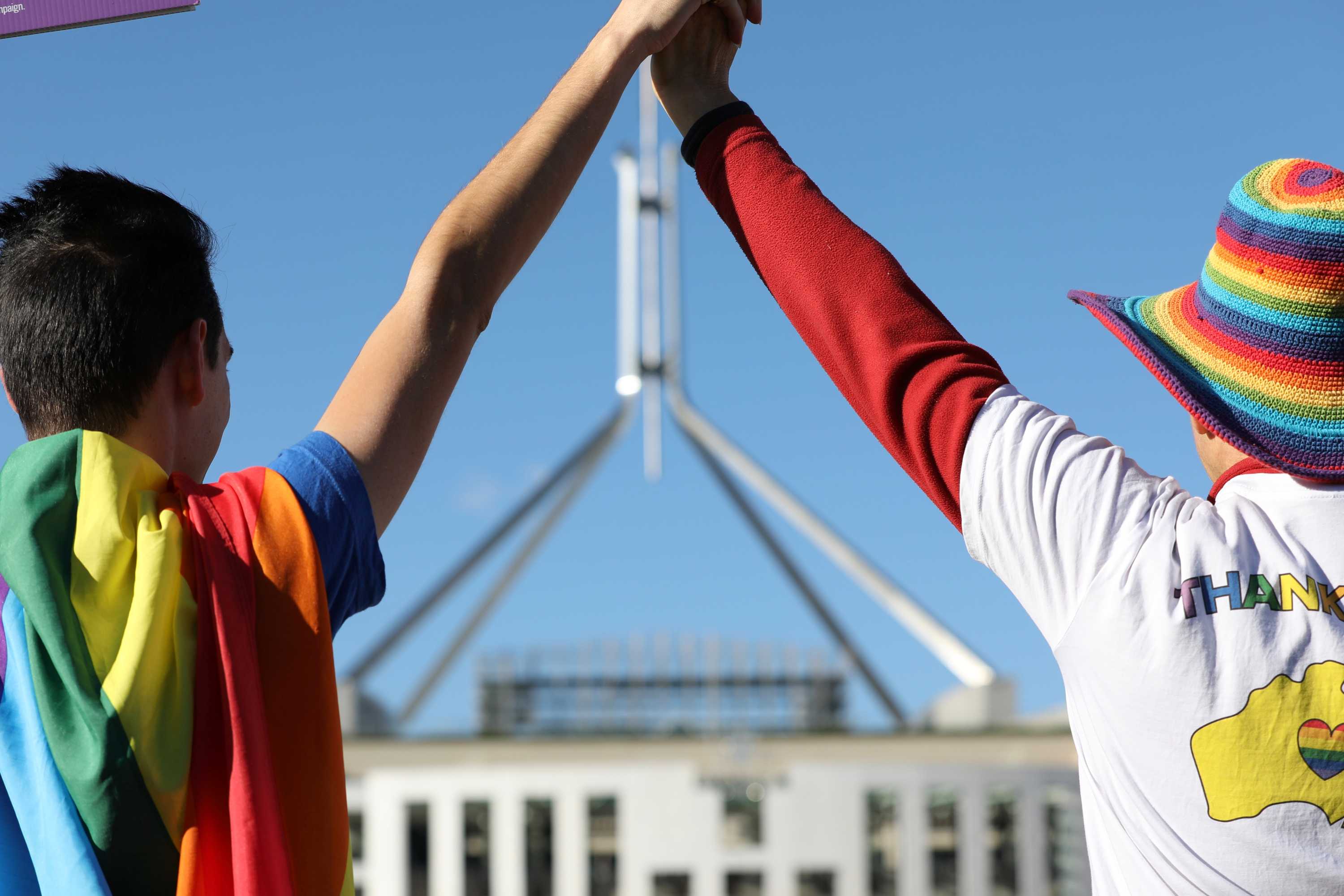 A person draped in a rainbow flag and another wearing a rainbow hat and a shirt that says "THANK YOU" outside Parliament