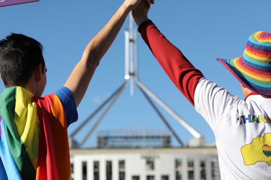 A person draped in a rainbow flag and another wearing a rainbow hat and a shirt that says "THANK YOU" outside Parliament