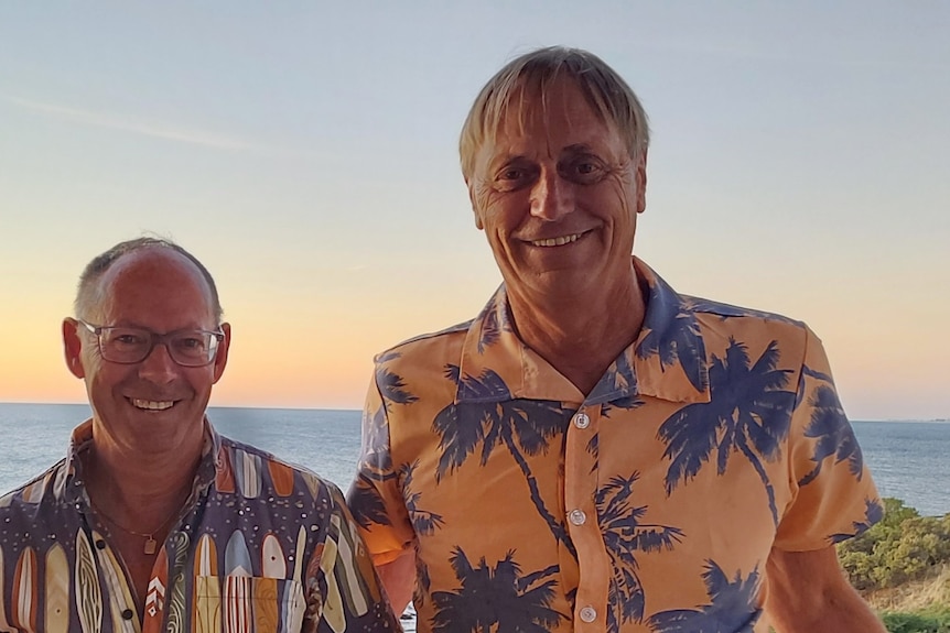 Двое мужчин улыбаются в камеру: у более низкого мужчины очки и лысеющая голова, у более высокого мужчины короткие светлые волосы.  Они носят гавайские рубашки.