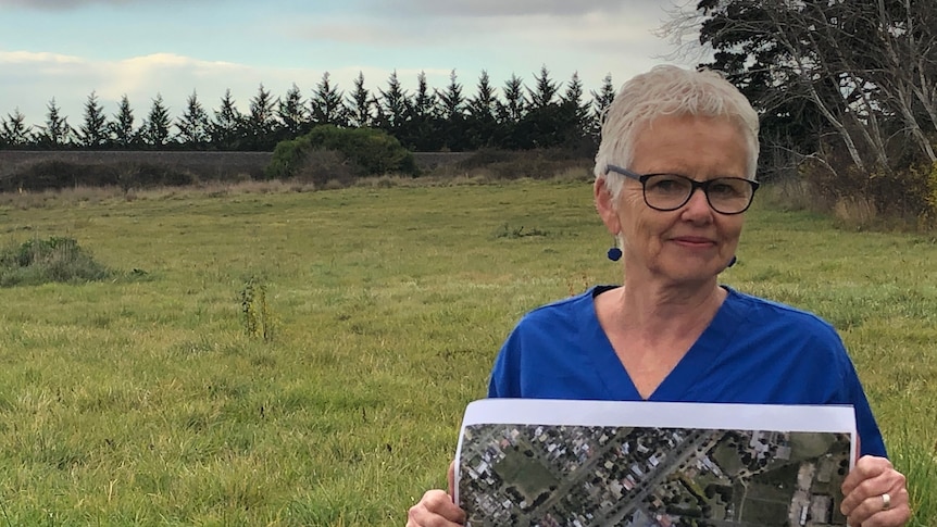 A lady with short white hair and black think-rimmed glasses holds a map of the proposed development in front of vacant land.