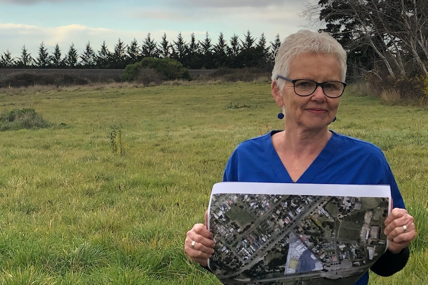 A lady with short white hair and black think-rimmed glasses holds a map of the proposed development in front of vacant land.