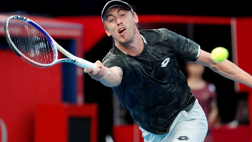 John Millman stretches with his racquet as he looks towards the ball wearing a black t-shirt and white shorts