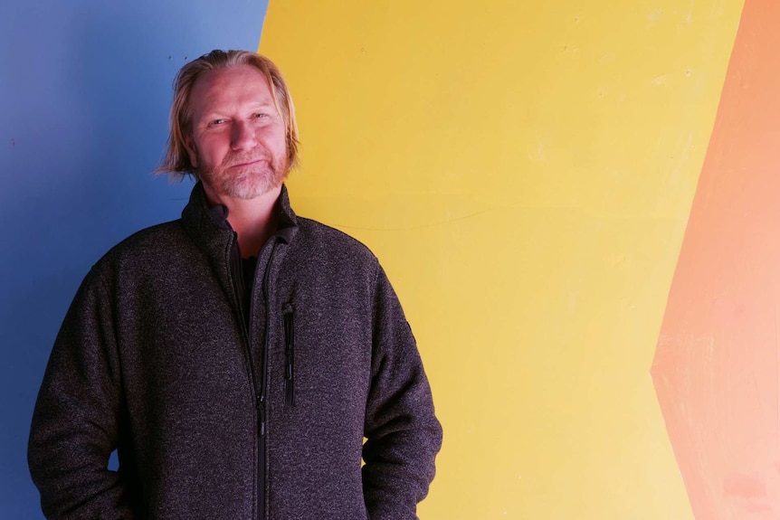 Medium close up photograph of a man with shoulder length hair and a blue jacket facing the camera