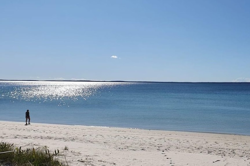 A figure stands on a beach in the middle of the day.