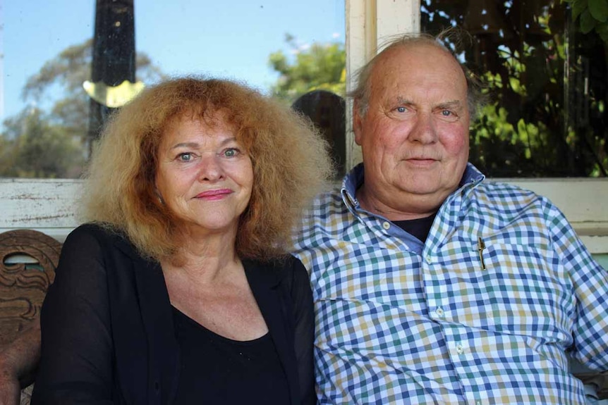 A couple aged in their 70's sit on a bench underneath a verandah at their farm.