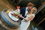A man and a woman on their wedding day, kissing while cutting a stack of cheese.