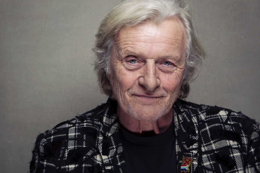 An aged Rutger Hauer smiles at the camera, wearing a black-and-white tartan blazer.