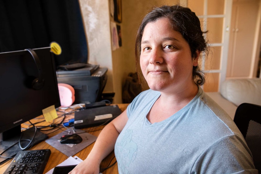 A young woman sits at a desk inside a house looking at the camera.