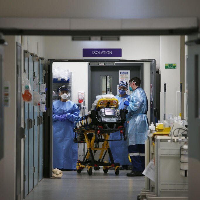 A corridor in a hospital with a patient on a bed surrounded by three medical staff in blue gowns and masks.